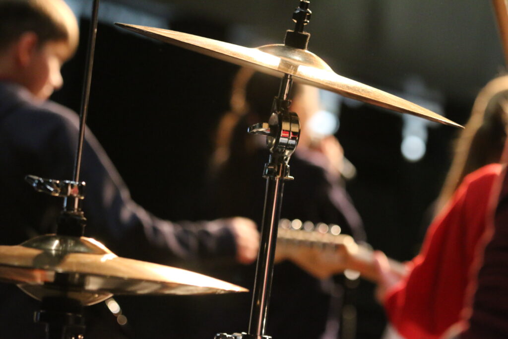 Photo shows a vocalist in the background with two guitar players and the cymbols in focus.