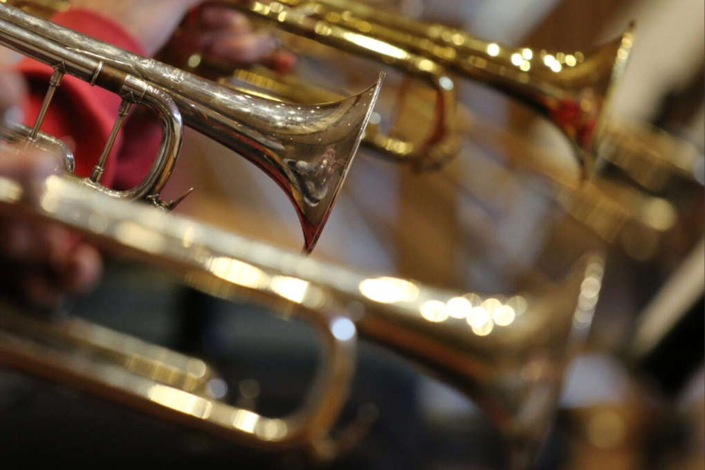 A row of shiny brass trumpets being played, shown close‑up with the focus on the instruments’ bells and valves.